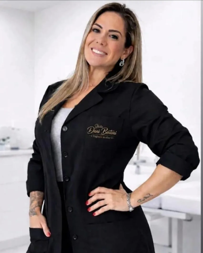 Dani Bertoni sorrindo, de jaleco preto com o logo bordado em dourado